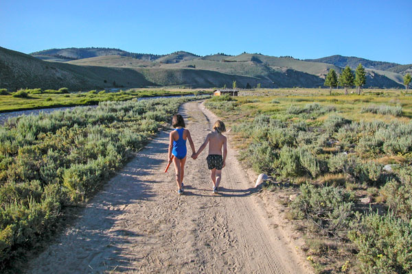 Two young children walking hand-in-hand down a rural road with mountains in the background